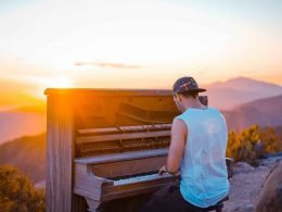 A man plays on a worn down upright piano on a hillside as the sun sets over the landscape.
