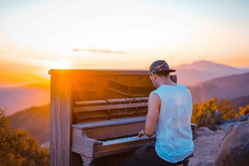 A man plays on a worn down upright piano on a hillside as the sun sets over the landscape.