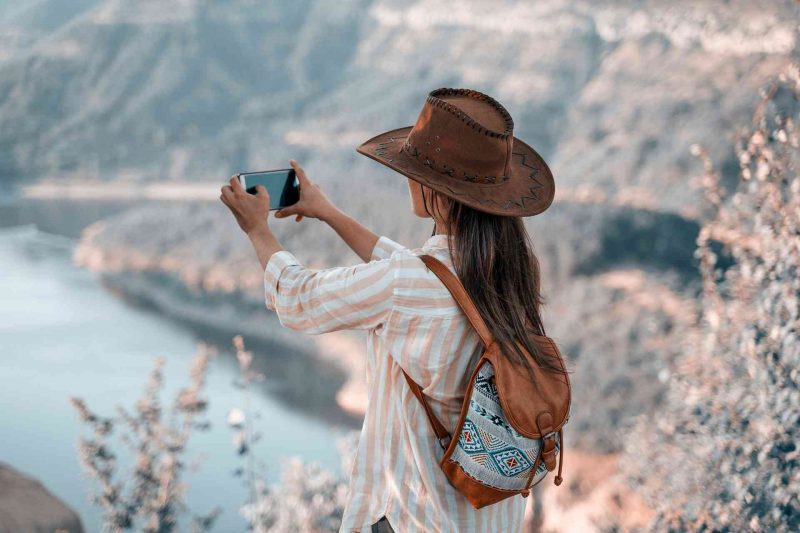 A woman captures a breathtaking view of a lake from high up in the mountains using her mobile phone.