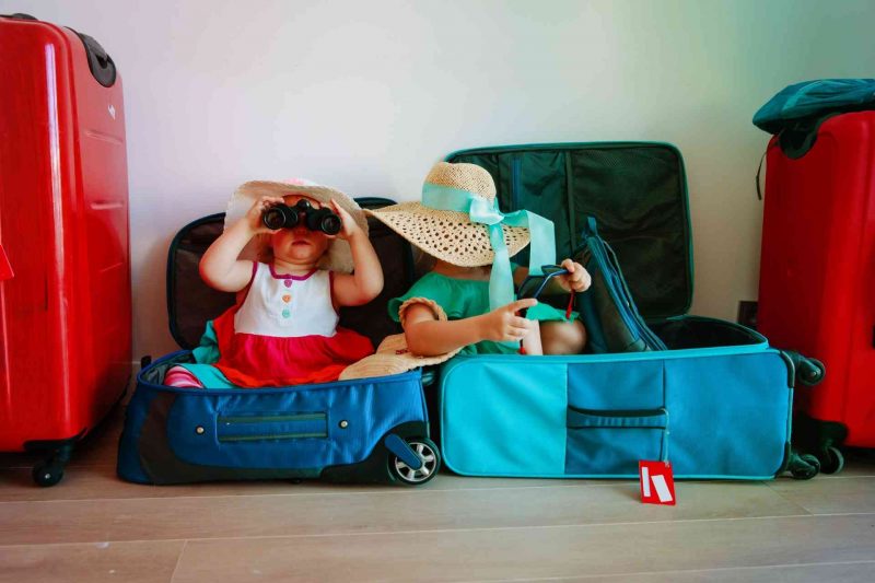 Two little girls sitting in suitcases playing with the packed hats and binoculars.