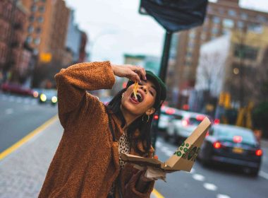A woman eating toppings and cheese from a New York style pizza as she walks along a city street.