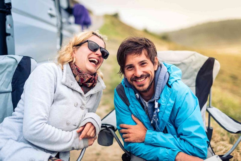 A happy couple smiling and laughing as they sit in camp chairs outside their RV.