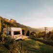 A stationary travel trailer parked ona hillside with a terrace attached overlooking the sunset.