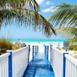 Walking down a blue and white board walk to a white sandy beach in the tropics.