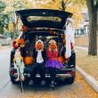 Two kids dressed in Halloween costumes sitting in the decorated trunk of an SUV parked along a street holding pumpkins up to their faces.