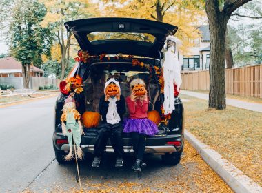 Two kids dressed in Halloween costumes sitting in the decorated trunk of an SUV parked along a street holding pumpkins up to their faces.