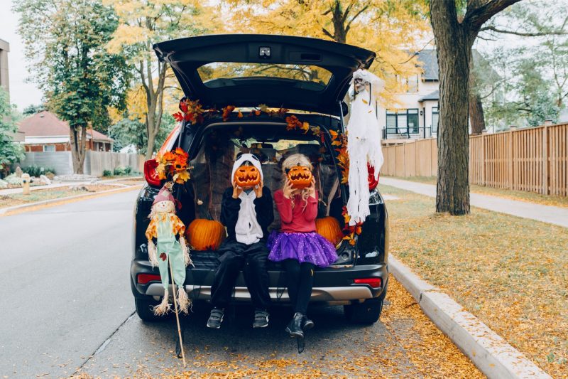 Two kids dressed in Halloween costumes sitting in the decorated trunk of an SUV parked along a street holding pumpkins up to their faces.