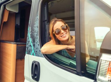 A woman leans out the window of the passenger seat in the cab of a Class B RV.