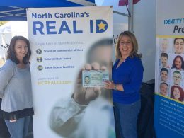 Two women stand in from of a REAL ID banner and one holds up her new Star ID.