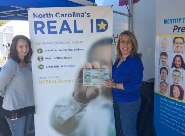 Two women stand in from of a REAL ID banner and one holds up her new Star ID.