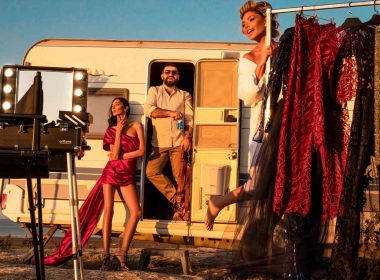 Three celebrities pose with racks of fashion clothes and lights outside of a travel trailer.