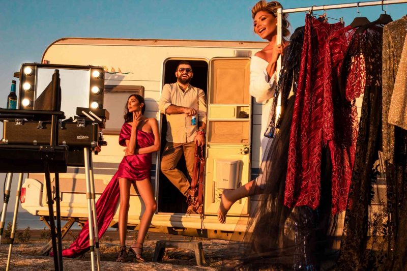 Three celebrities pose with racks of fashion clothes and lights outside of a travel trailer.