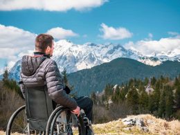 A man with a winter jacket looks out over majestic mountains from his wheelchair.