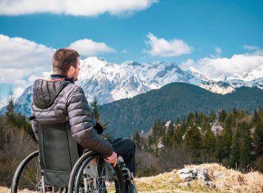 A man with a winter jacket looks out over majestic mountains from his wheelchair.