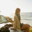 A woman in a cardigan and summer dress relaxes on the hood of her car by the beach and the sun begins to set.