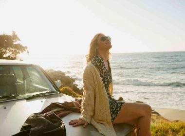 A woman in a cardigan and summer dress relaxes on the hood of her car by the beach and the sun begins to set.