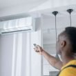 A man uses a remote to turn on his mini split air conditioner mounted high up on the wall of his home.