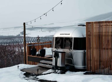 A shiny aluminum Airstream travel trailer parked at the top of a snowy hill overlooking the valley below.