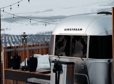 A shiny aluminum Airstream travel trailer parked at the top of a snowy hill overlooking the valley below.