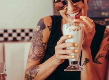 Young woman with shades on sits in a retro booth drinking a creamy soda from a straw.