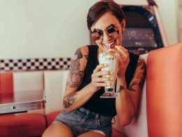 Young woman with shades on sits in a retro booth drinking a creamy soda from a straw.