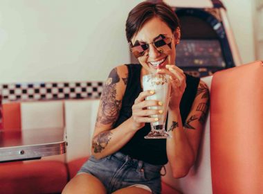 Young woman with shades on sits in a retro booth drinking a creamy soda from a straw.