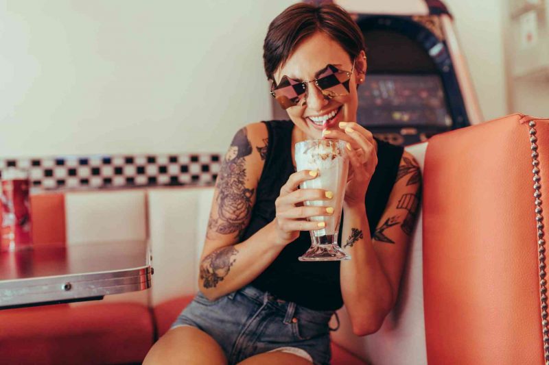 Young woman with shades on sits in a retro booth drinking a creamy soda from a straw.