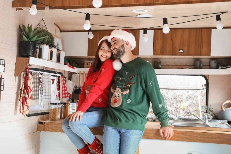 A couple adorned in Christmas sweaters and Santa hats having fun in their RV.