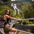 A solo female traveler with a backpack leans on a railing in front of a waterfall in a rainforest.