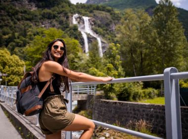 A solo female traveler with a backpack leans on a railing in front of a waterfall in a rainforest.