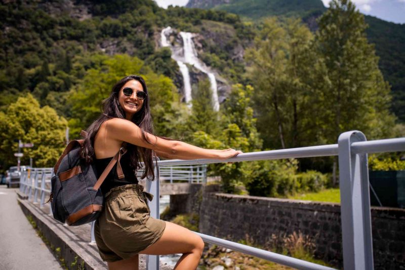 A solo female traveler with a backpack leans on a railing in front of a waterfall in a rainforest.