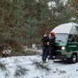 A couple in winter jackets and boots standing outside of their camper van parked on a snow covered road in the woods.