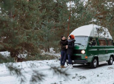 A couple in winter jackets and boots standing outside of their camper van parked on a snow covered road in the woods.