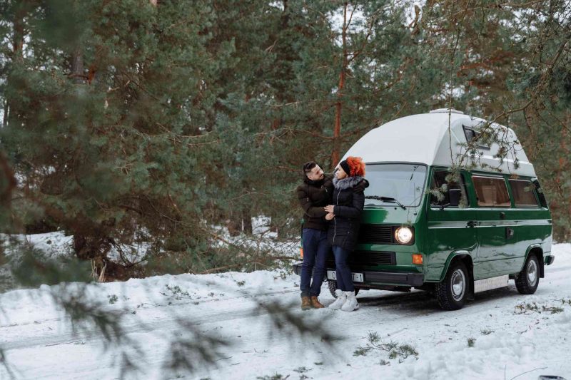 A couple in winter jackets and boots standing outside of their camper van parked on a snow covered road in the woods.