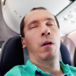 Man sleeping on a plane wrong in a teal shirt and no neck pillow.