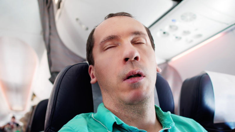 Man sleeping on a plane wrong in a teal shirt and no neck pillow.