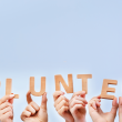 The word volunteer being spelled out with wooden blocks that are held by individual hands with a blue background.