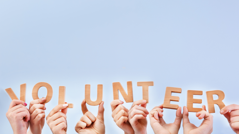 The word volunteer being spelled out with wooden blocks that are held by individual hands with a blue background.