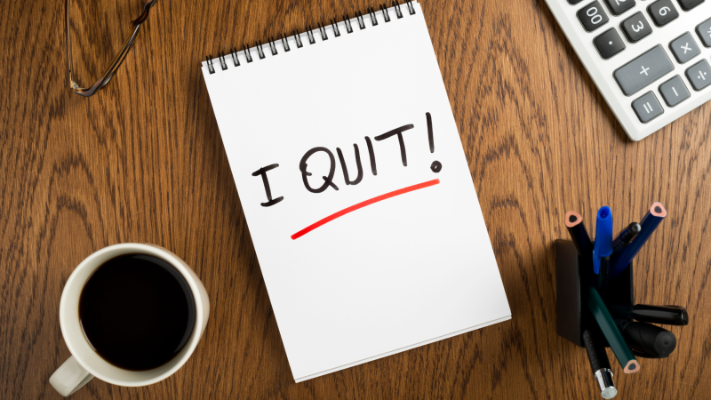 "I QUIT!" wirtten on a notepad on a desk between a mug of coffe and pens.
