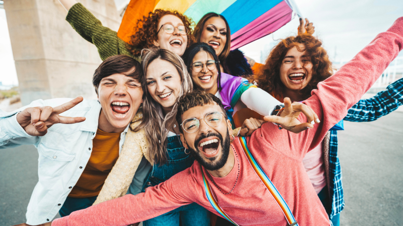 A group of seven people happily taking a photo together with an lgbtq+ flag in the background.