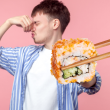 A man plugging their nose as they stand in front of a pink backdrop as a piece of sushi is being held in front of him with chopsticks.