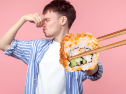 A man plugging their nose as they stand in front of a pink backdrop as a piece of sushi is being held in front of him with chopsticks.