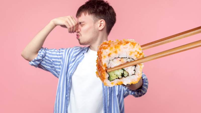 A man plugging their nose as they stand in front of a pink backdrop as a piece of sushi is being held in front of him with chopsticks.