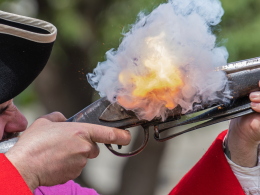 A close up of a man in red reenacting as he shoots off a gun.