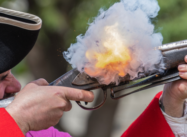 A close up of a man in red reenacting as he shoots off a gun.