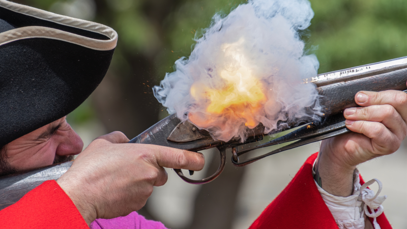 A close up of a man in red reenacting as he shoots off a gun.