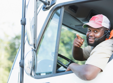 A person smiling and pointing at the camera as they hang out of their truck window.