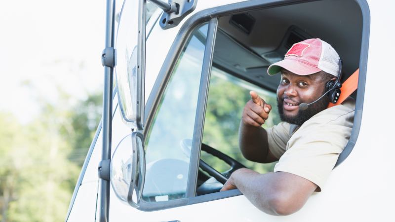 A person smiling and pointing at the camera as they hang out of their truck window.