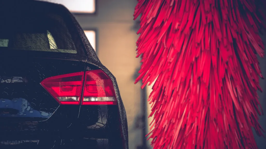 A black car driving through an automated car wash as a red brush sits idle beside it. 