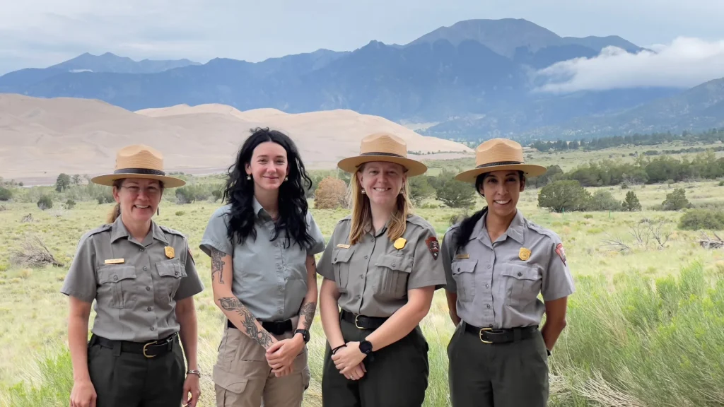 Four female park rangers of the Great Sand Dunes National Park stand side by side with sand dunes in the background and mountains.